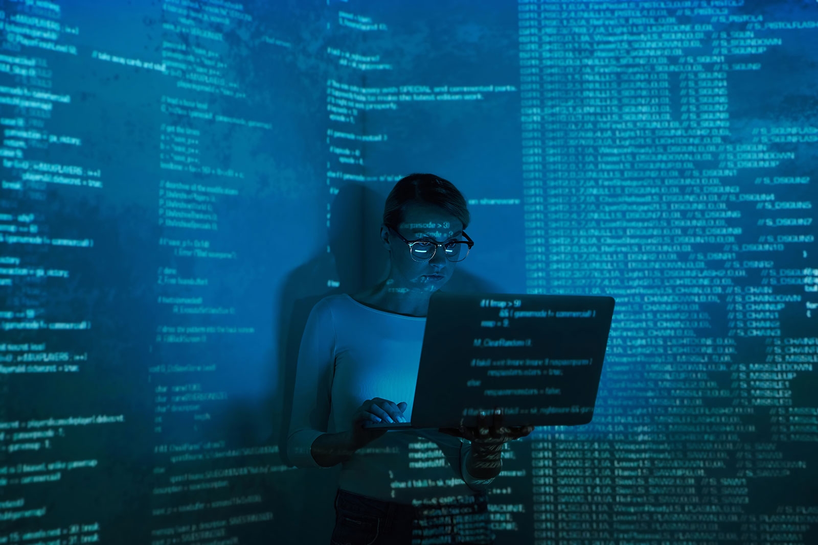 Woman in glasses holding an open laptop, standing against a wall with lines of code projected across her