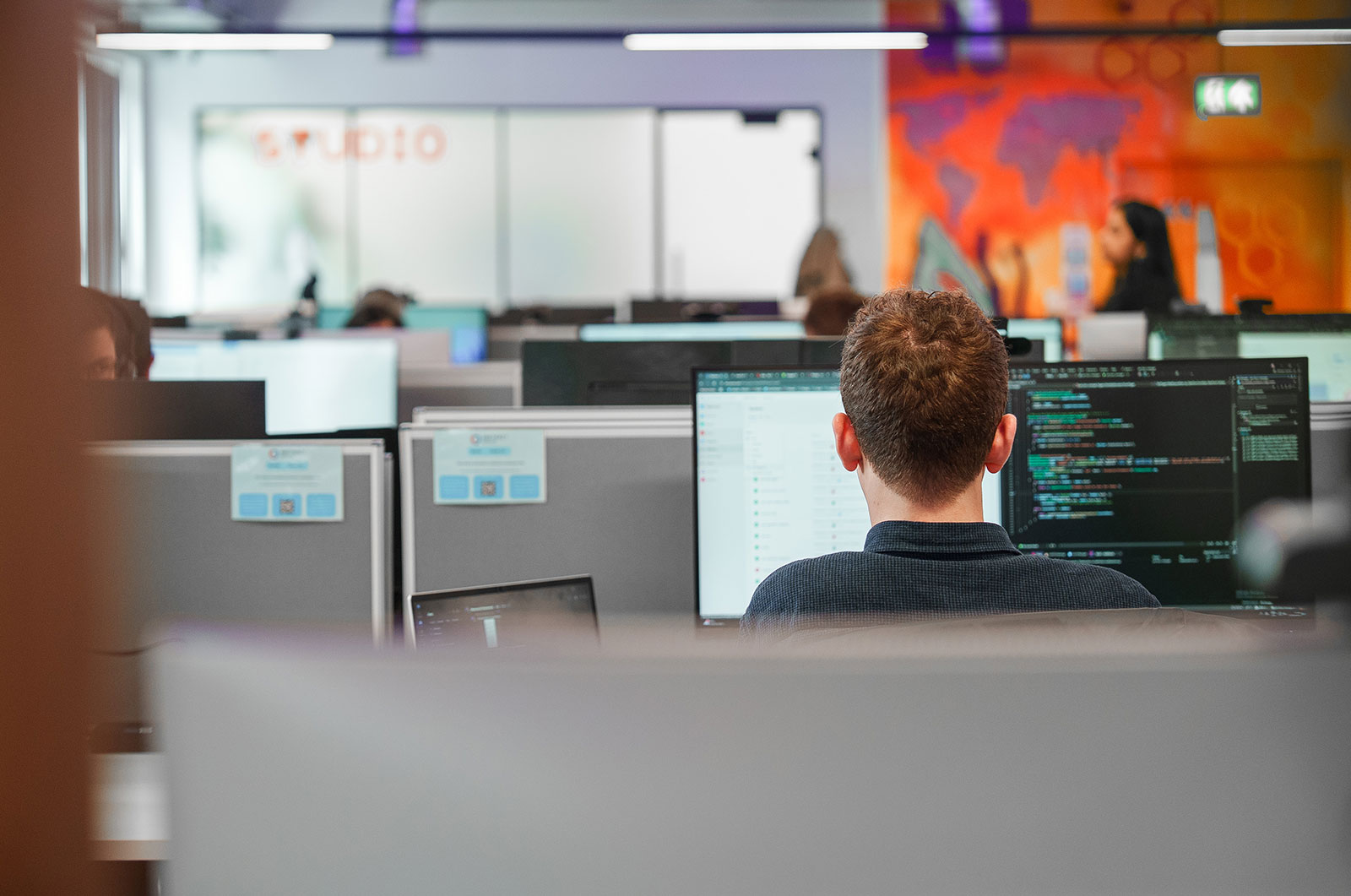 Engineer seen from behind, working on code across two monitors in our open-plan office, with a colourful world map mural beyond