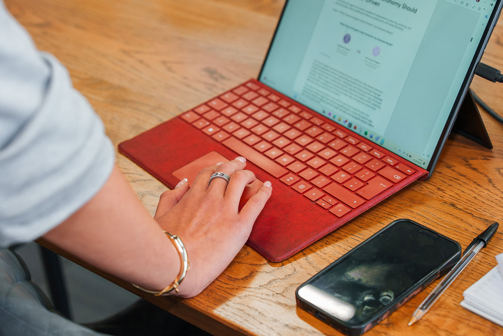Close-up of a hand using the trackpad of a tablet with a red keyboard cover, a document open on screen, on a wooden desk