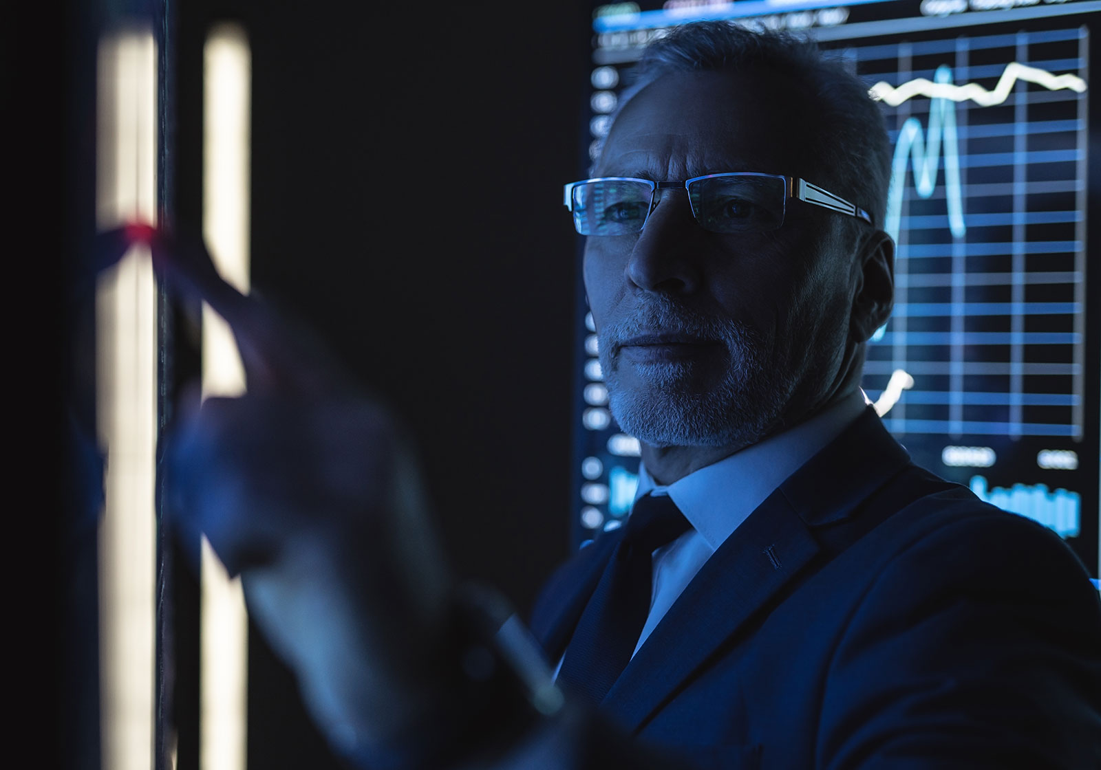A man in a suit and glasses, lit by blue light, reaching towards a screen with a data chart behind him
