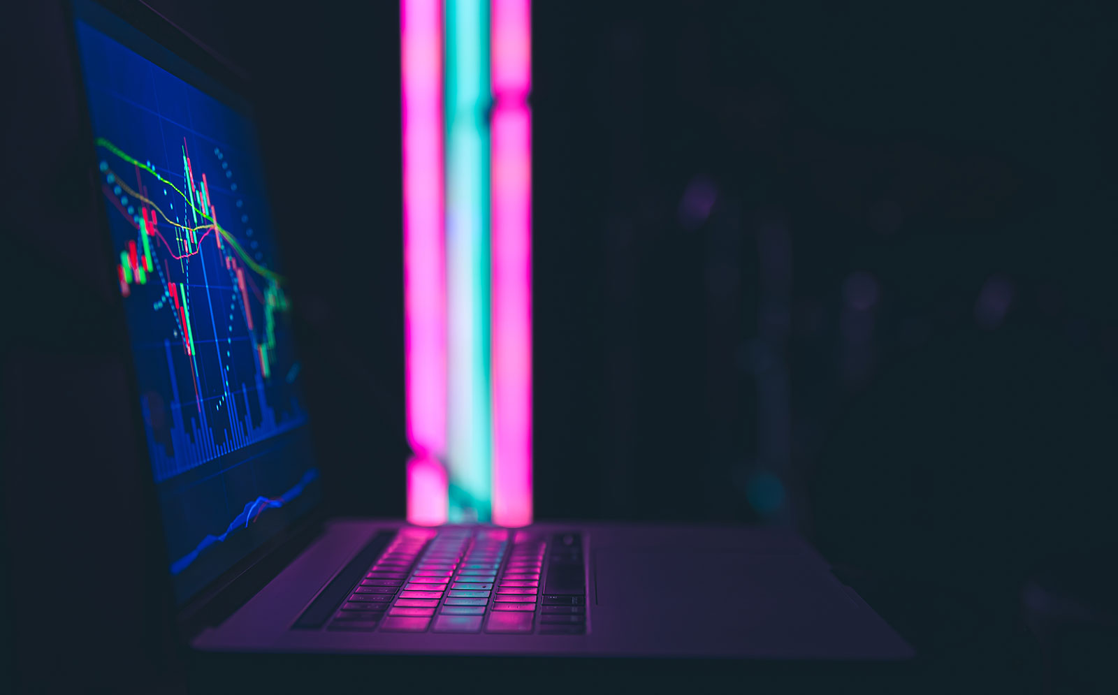 Laptop in a dark room displaying a financial candlestick chart, its keyboard lit by pink and teal neon light